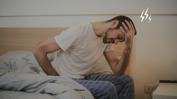 Ein Mann mit braunen Haaren und Bart sitzt in weißem T-Shirt und blau karierter Stoffhose auf der Kante seines Bettes mit gestreifter Bettwäsche und sützt seinen Kopf in seine linke Hand, während er die Augen zu hat und sein Gesicht leicht verzieht.