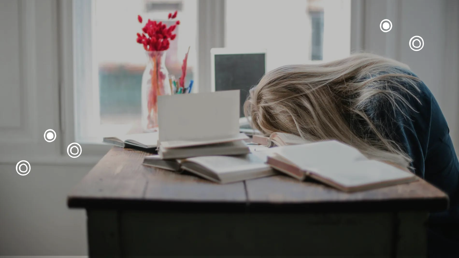 Eine Frau mit blonden Haaren sitzt vor einem Stapel aufgeschlagener Bücher und einem Laptop an einem Holztisch mit roten Blumen und liegt mit ihrem Gesicht auf einem der Bücher.