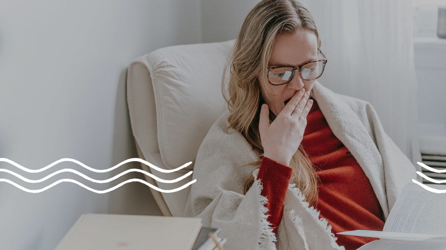 Eine blonde Frau mit Brille im roten Pullover und einer weißen Decke um die Schultern sitzt auf einem Sessel und gähnt, während sie ein Buch liest.