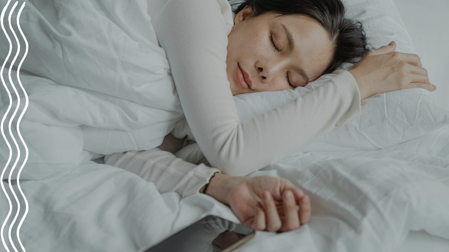 Eine Frau mit schwarzen Haaren und weißem Langarmshirt liegt auf ihrer linken Seite in einem Bett mit weißer Bettwäsche und schläft. Neben ihr liegt ein schwarzes Handy.
