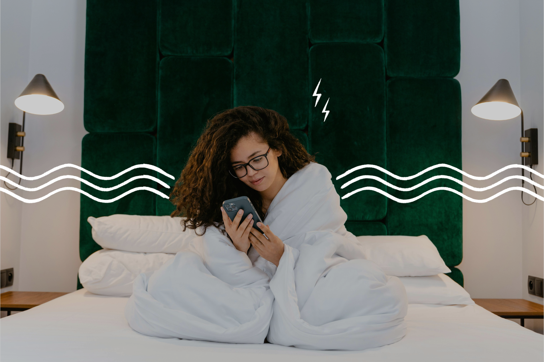 Eine junge Frau mit Brille sitzt in eine Decke gehüllt auf einem Bett und schaut auf ihr Handy