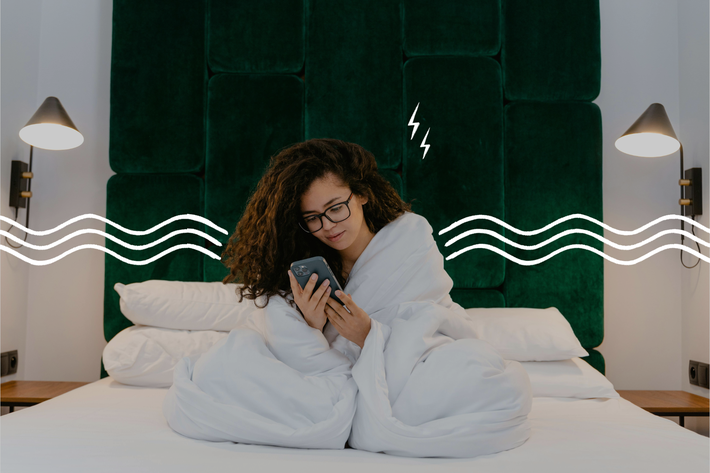 Eine junge Frau mit Brille sitzt in eine Decke gehüllt auf einem Bett und schaut auf ihr Handy