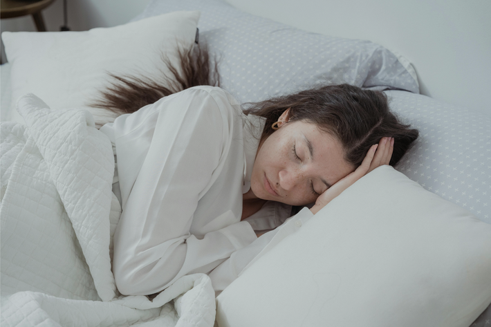 Junge Frau mit geschlossen Augen schläft in Seitenlage, trägt ein weißes Langarmshirt und liegt in weißer Bettwäsche – Symbolbild für Atemübungen zum Einschlafen.