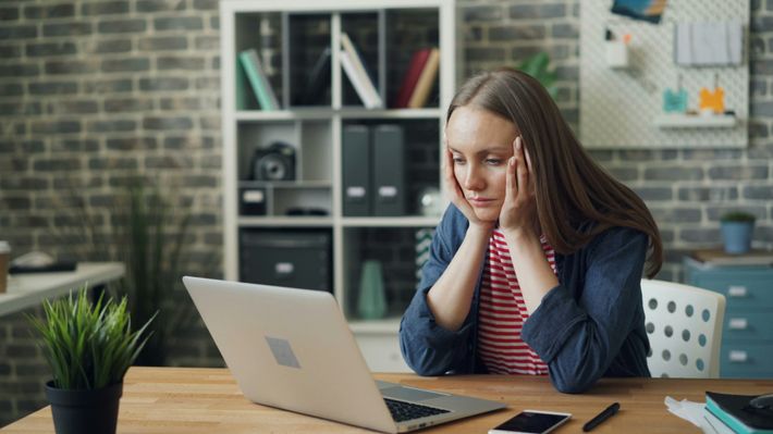 Eine Frau mit langen Haaren, rot-weiß-gestreiftem Oberteil und blauem Hemd sitzt auf einem weißen Stuhl mit Löchern an einem Holzschreibtisch und starrt müde auf ihren Laptop, mit ihrem Gesicht in die Hände gestützt. Auf dem Tisch steht eine Pflanze, ihr Hand, Stift und Notizbücher. Im Hintergrund sieht man ein Regal mit Ordnern, eine Pinnwand, einen Schrank und eine gemauerte Wand.