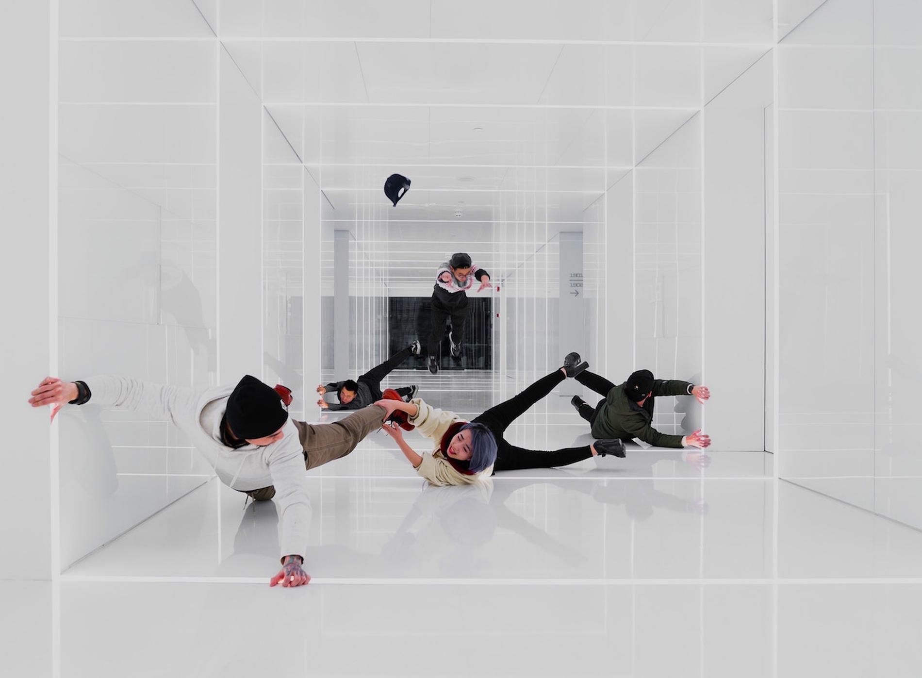 Vier Personen befinden sich in einem futuristisch wirkenden, komplett weißen Raum mit spiegelnden Oberflächen. Sie scheinen in der Luft zu schweben oder zu stürzen, als ob die Schwerkraft aufgehoben wäre. Ihre Körper sind in ungewöhnlichen Positionen, was eine surreale oder inszenierte Szene vermuten lässt. Ein Baseball-Cap schwebt ebenfalls durch den Raum. Die Szene erinnert an eine optische Illusion oder eine kreative Tanz- oder Kunstinstallation.