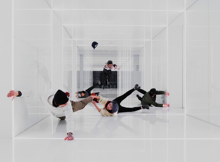 Vier Personen befinden sich in einem futuristisch wirkenden, komplett weißen Raum mit spiegelnden Oberflächen. Sie scheinen in der Luft zu schweben oder zu stürzen, als ob die Schwerkraft aufgehoben wäre. Ihre Körper sind in ungewöhnlichen Positionen, was eine surreale oder inszenierte Szene vermuten lässt. Ein Baseball-Cap schwebt ebenfalls durch den Raum. Die Szene erinnert an eine optische Illusion oder eine kreative Tanz- oder Kunstinstallation.