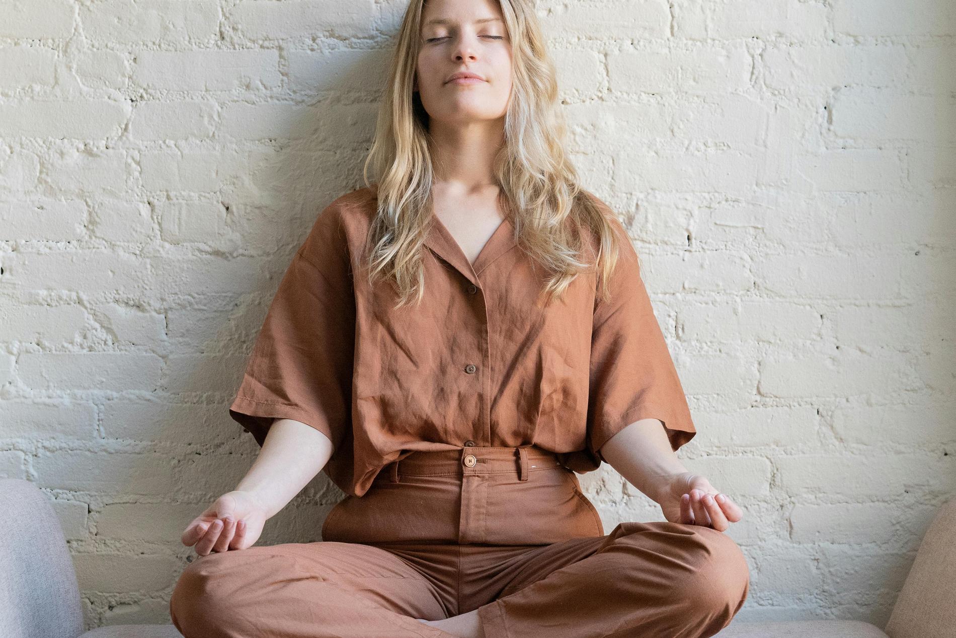 Eine blonde Frau mit braunem Leinenoberteil und brauner Hose sitzt mit geschlossenen Augen im Schneidersitzt vor einer weißen Mauerwand mit ihren Händen auf den Knien in Meditationshaltung und wirkt entspannt.