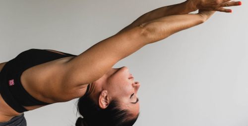 Frau mit braunen Haaren und einem Sportoberteil, die eine Rückbeuge macht und die Arme über ihren Kopf ausstreckt.