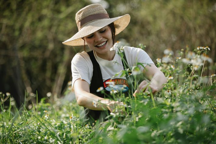 Eine junge Frau mit hellem Strohhut und T-Shirt kniet in einem Garten und pflückt oder pflegt weiße Gänseblümchen-ähnliche Blumen, während sie fröhlich lächelt und dankbar aussieht.