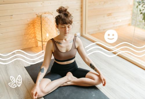 Frau mit braunen Haaren, Sportoutfit und Tattoos, die auf einer Yogamatte neben einem Fenster meditiert