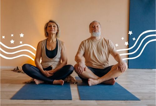 Eine Frau und einem Mann mit grauem Bart im mittleren Alter, die auf blauen Yogamatten sitzen und die Augen geschlossen haben.