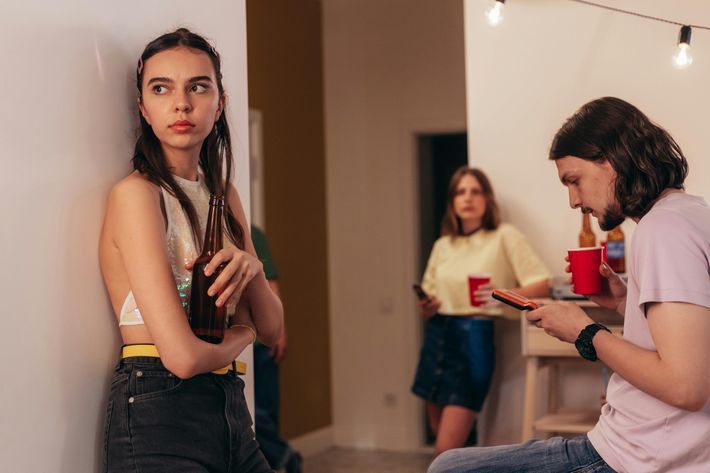 Zwei Frauen und ein Mann sind in einem Zimmer: Eine Frau steht links gegen eine Wand gelehnt mit einer braunen Glasflasche in der Hand, einem glitzerndem Oberteil und grauer Jeans, während sie zur Seite schaut. Der Mann rechts sitzt und schau auf ein Gerät, während er einen roten Becher in seiner rechten Hand hält. Die andere Frau ist verschwommen und steht im Hintergrund mit rotem Becher in der Hand, einem Jeansrock und einem gelben T-Shirt.