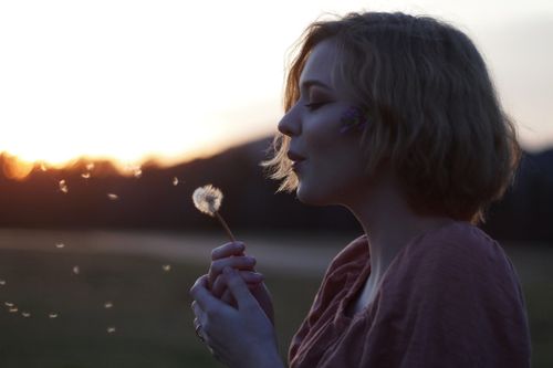 Profil-Aufnahme einer Frau im Sonnenuntergangslicht, die eine Pusteblume bläst.