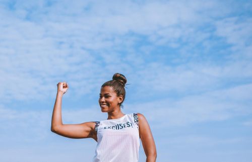 Frau in einem Sportoberteil, die lachend die Faust in den blauen Himmel reckt.