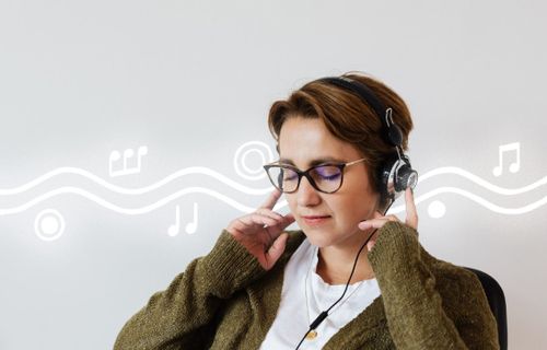 Frau mit kurzen rötlichen Haaren und Brille, die mit geschlossenen Augen Kopfhörer trägt und der Musik lauscht.