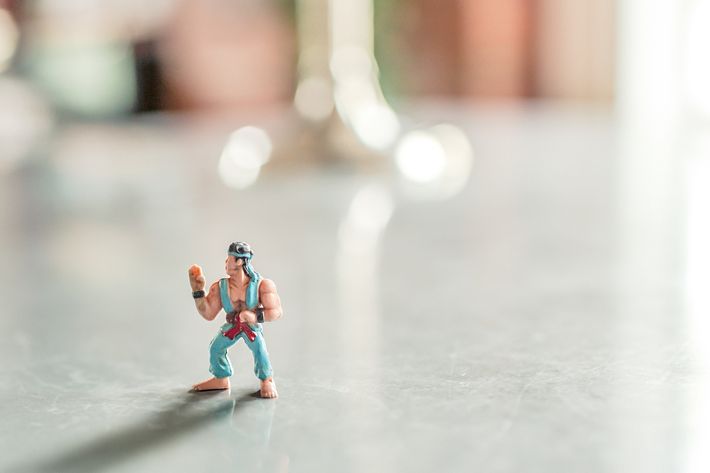 Eine Spielfigur steht auf einem grauen Untergrund, trägt blaue Kleidung und ein blaues Stirnband und hebt die rechte Hand. Der Hintergrund ist verschwommen.