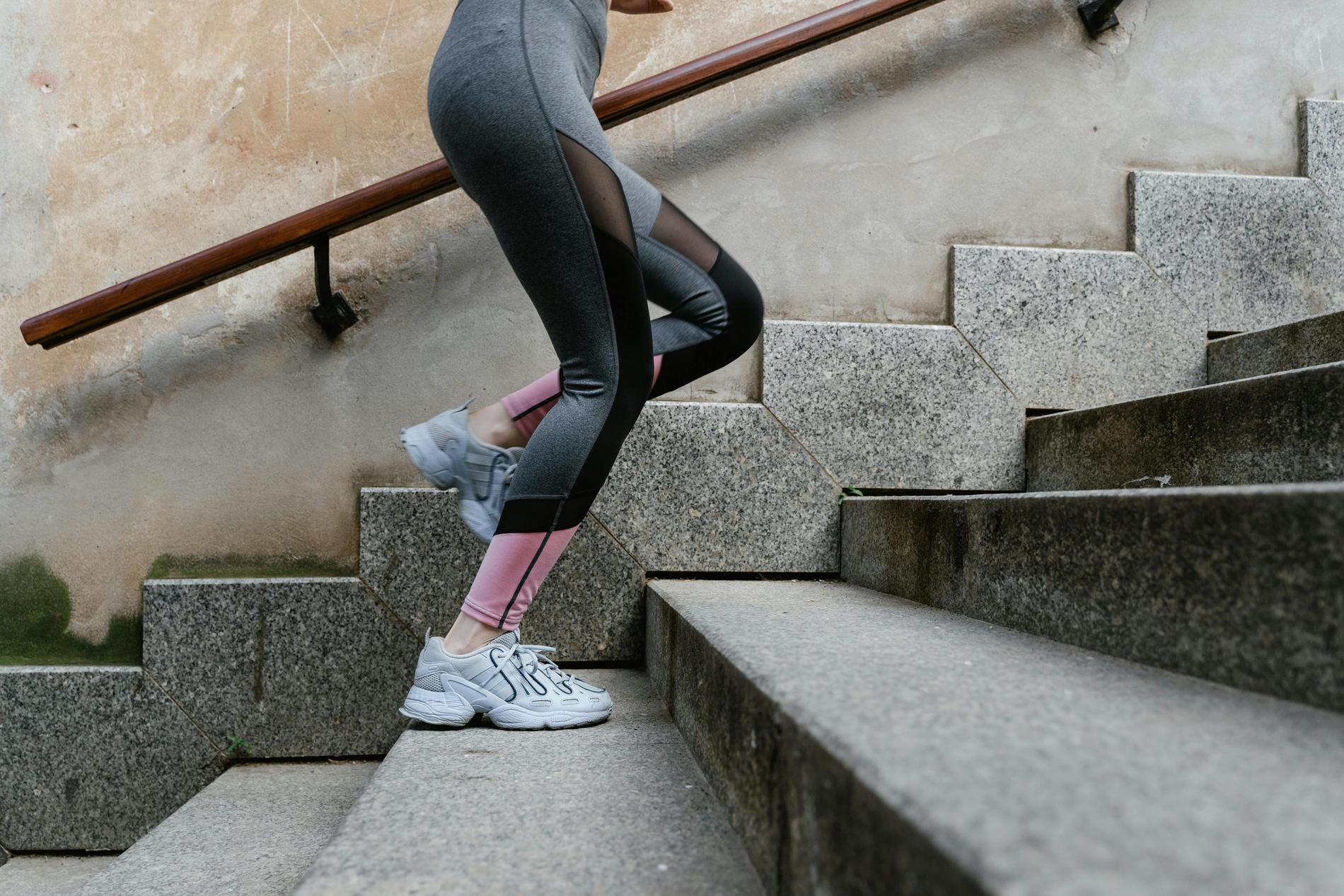 Man sieht die Beine einer Person in grau-schwarzer Sportleggings mit weißen Sportschuhen von der Seite, wie sie eine graue Treppe hochläuft.