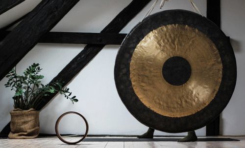 Großer Gong in den Farben schwarz und gold vor einer weißen Wand und neben einer Zimmerpflanze in einem Topf aus Jute.