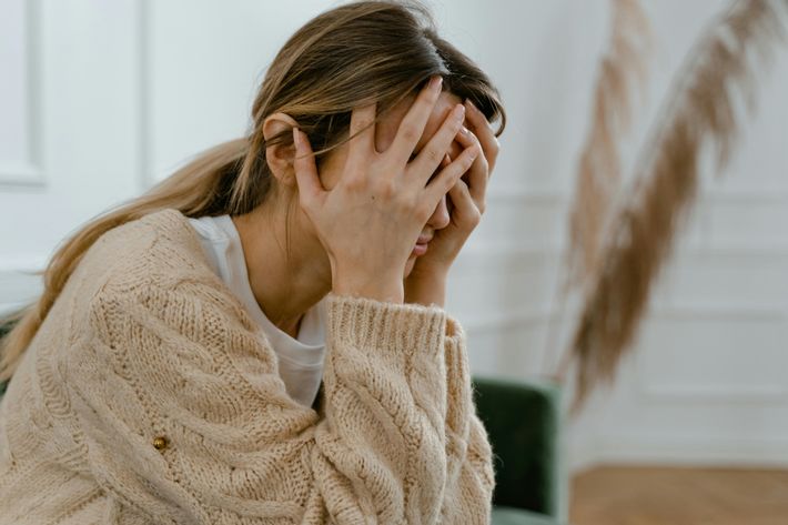 Eine Frau mit beige gemustertem Pullover und blonden, langen Haaren in einem Zopf vergräbt ihr Gesicht in ihren Händen. Der Hintergrund ist verschwommen.
