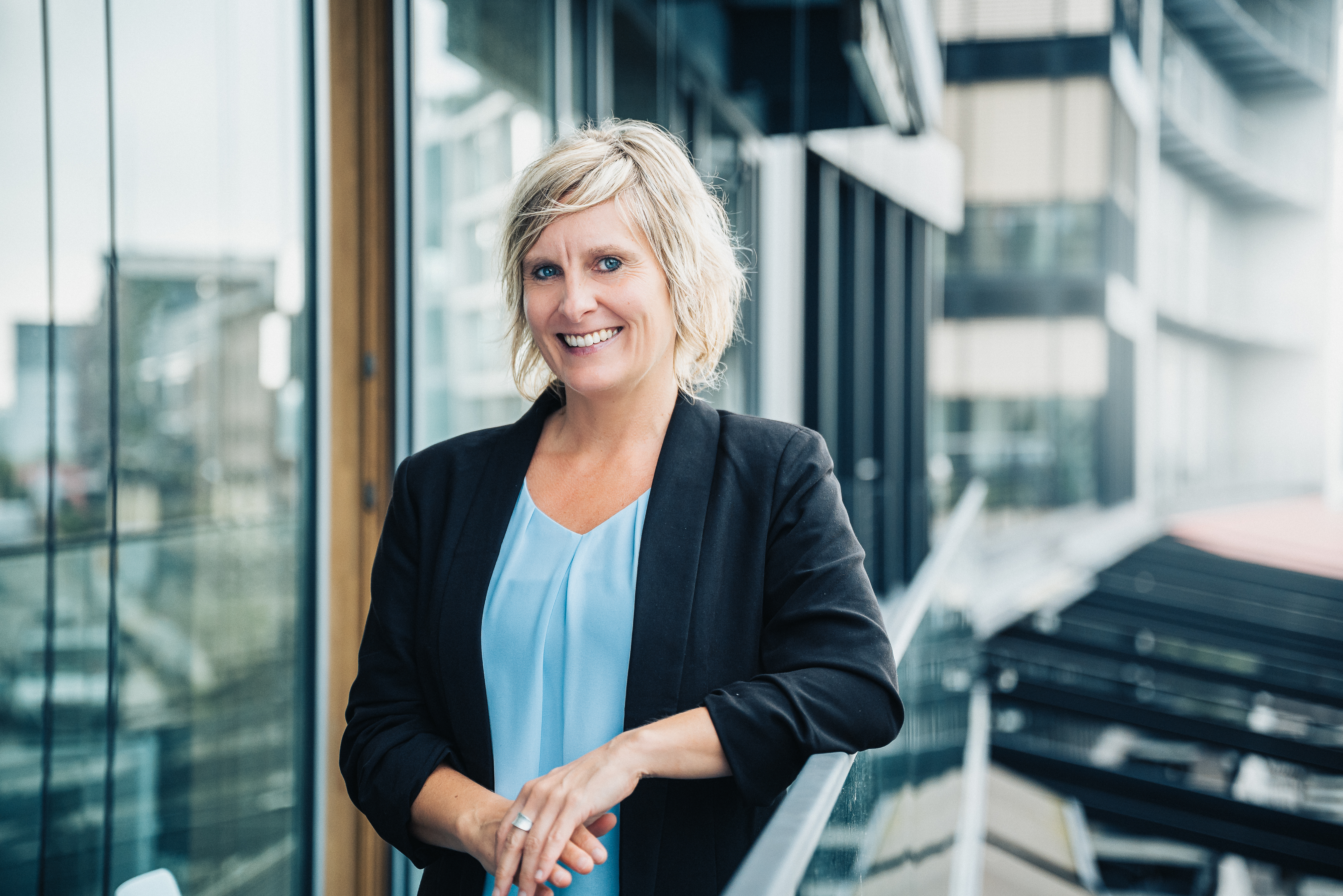Systemische Business & Personal Coachin Verena Arnhold schaut mit schwarzen Blazer und hellblauem Oberteil mit ihrem linken Arm auf einem Geländer gestützt lachend in die Kamera. Im Hintergrund sieht man verschwommen Fenster und Gebäude.