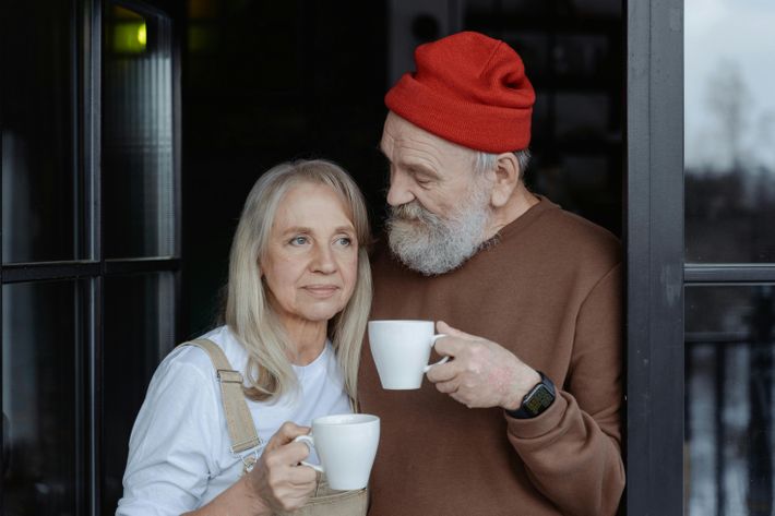 Eine ältere Frau und ein älterer Mann stehen Arm in Arm am Fenster mit je einer weißen Tasse in der Hand. Die Frau schaut in die Ferne, der Mann schaut sie an und trägt eine rote Mütze.