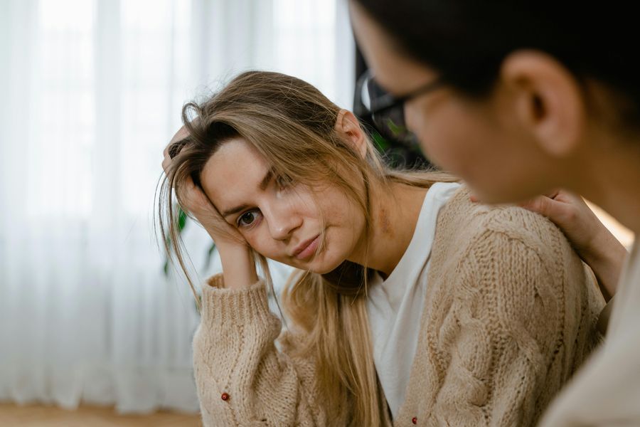 Eine blonde Frau mit weißem T-Shirt und beiger Strickjacke im Zopfmuster stützt ihren Kopf auf ihre rechte Hand, starrt nach vorne und sieht gestresst aus. Neben ihr sitzt eine braunhaarige Frau mit Brille und hat eine Hand auf die Schulter der blonden Frau gelegt. Im Hintergrund sind weiße Gardinen.