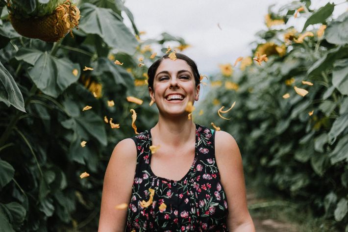 Eine Frau mit braunen Haaren und Blumenkleid steht im Grünen umringt von Sonnenblumen, deren gelbe Blütenblätter durch die Luft fliegen, während die Frau lacht.