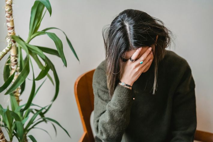 Eine Person mit dunklem, schulterlangem Haar sitzt auf einem Stuhl und hält sich mit der Hand das Gesicht, als ob sie gestresst oder ängstlich ist. Sie trägt einen dunklen Pullover und mehrere Ringe sowie ein Armband. Neben ihr steht eine grüne Zimmerpflanze.
