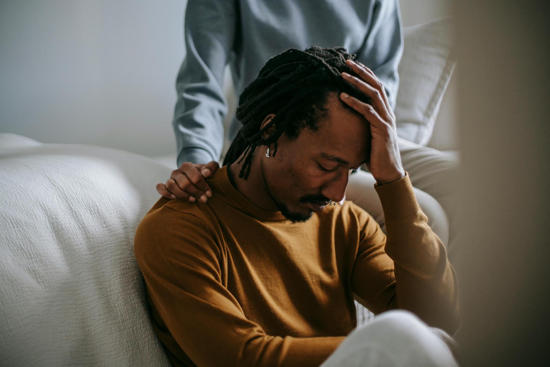 Ein junger Mann sitzt auf einem Sofa, stützt den Kopf mit seiner linken Hand ab und hat die Augen geschlossen. Man sieht, wie jemand hinter ihm steht und die Hand unterstützend auf seiner Schulter liegen hat.