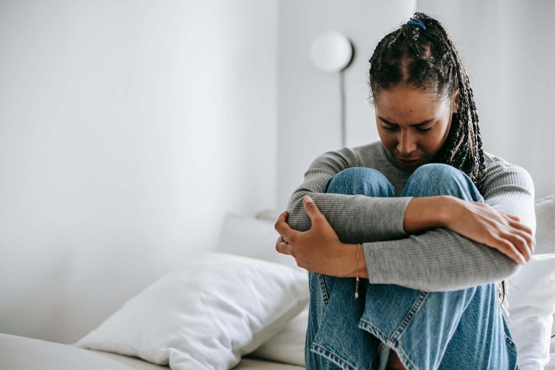 Eine Frau mit schwarzen Haaren im Zopf, grauem Langarmshirt und blauer Jeans sitzt mit angezogenen Beinen auf einem Bett mit weißer Bettwäsche und schaut deprimiert nach unten.