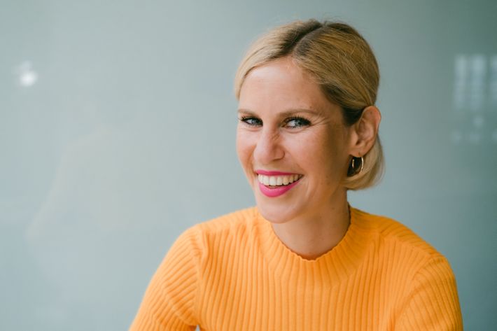 Dr. Eva Elisa Schneider im orangenen Pullover mit pinken Lippenstift vor einem grauen Hintergrund schaut und lacht in die Kamera.