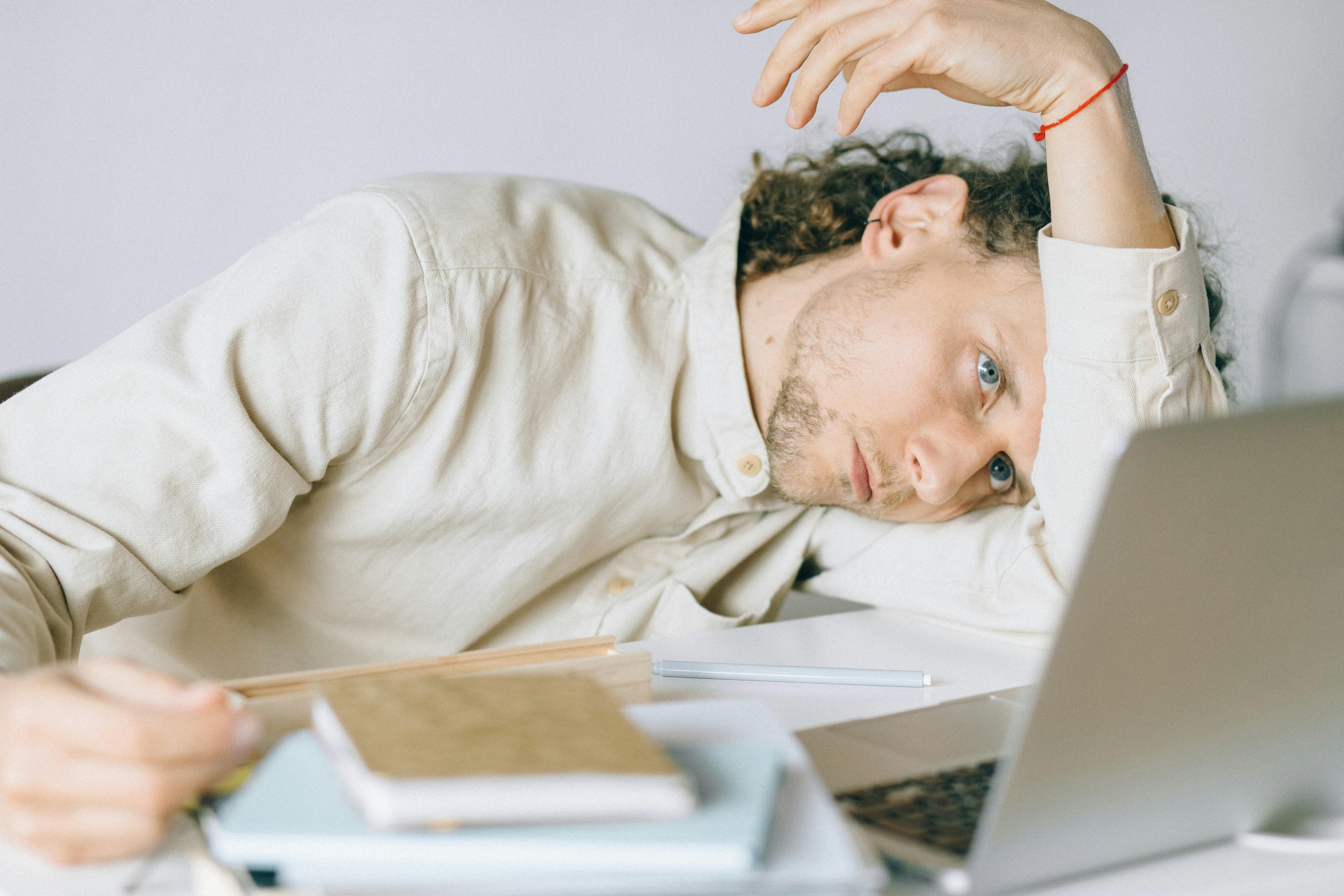 Ein junger Mann mit lockigem Haar liegt erschöpft auf einem Schreibtisch, den Kopf auf dem Arm abgestützt. Er trägt ein helles Hemd und blickt müde oder gedankenverloren zur Seite. Vor ihm liegen ein Laptop, Notizbücher, Stifte und weitere Arbeitsmaterialien. Die Szene vermittelt einen Eindruck von Erschöpfung oder Überforderung.