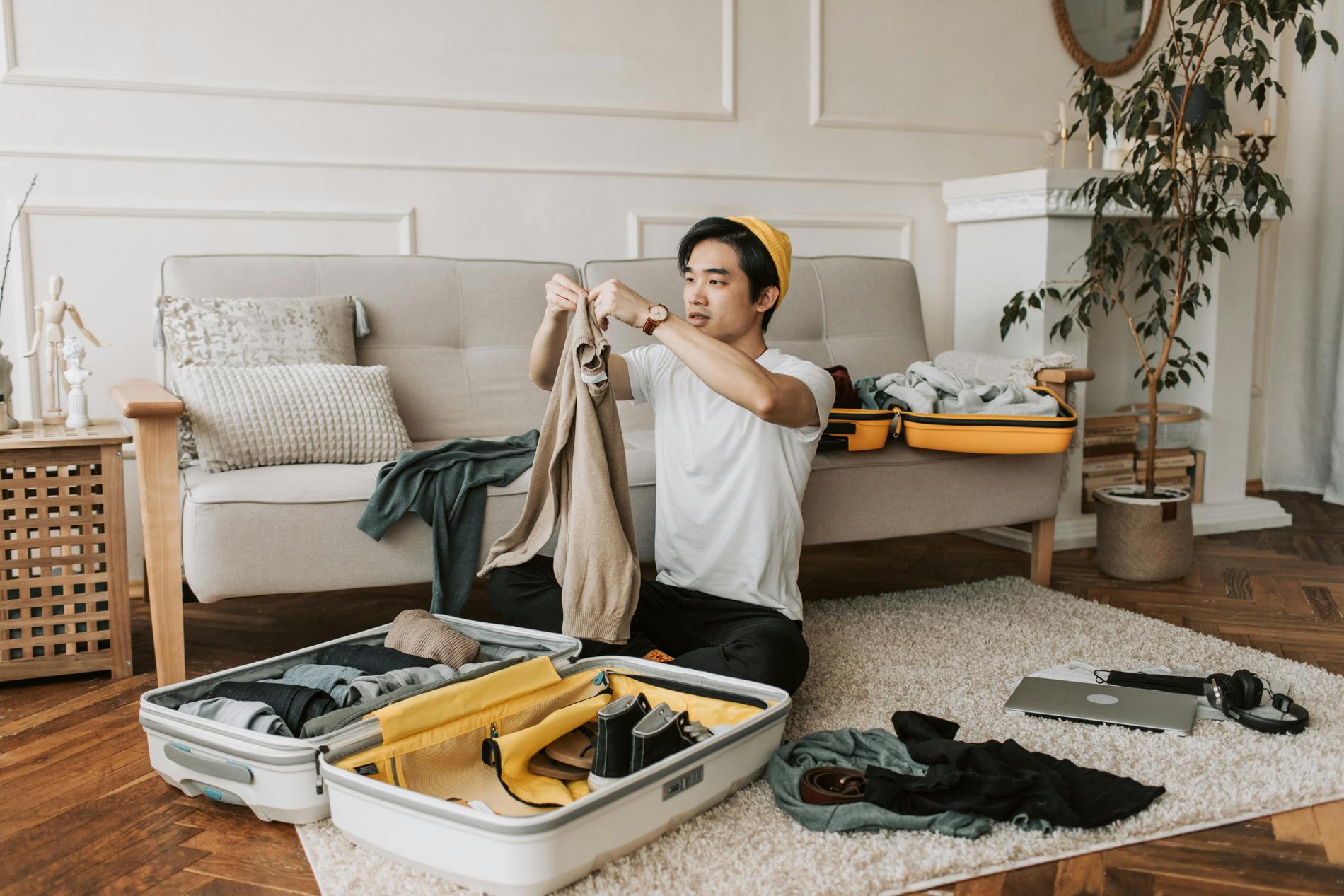 Ein Mann mit schwarzen Haaren mit gelber Mütze und weißem T-Shirt sitzt auf einem weißen Teppich und packt einen Koffer, der geöffnet vor ihm liegt. Um ihn herum liegen Klamotten, ein Laptop, Kopfhörer. Im Hintergrund sieht man ein helles Sofa, auf dem ein weiterer, kleiner Koffer liegt, eine Pflanze und Bücher.