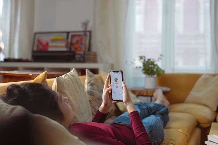 Eine Frau liegt barfuß und in Jeans und rotem Langarmshirt auf einem gelben Sofa und schaut auf ihr Handy, auf dem sie ein Tinder Dating Profil geöffnet hat.