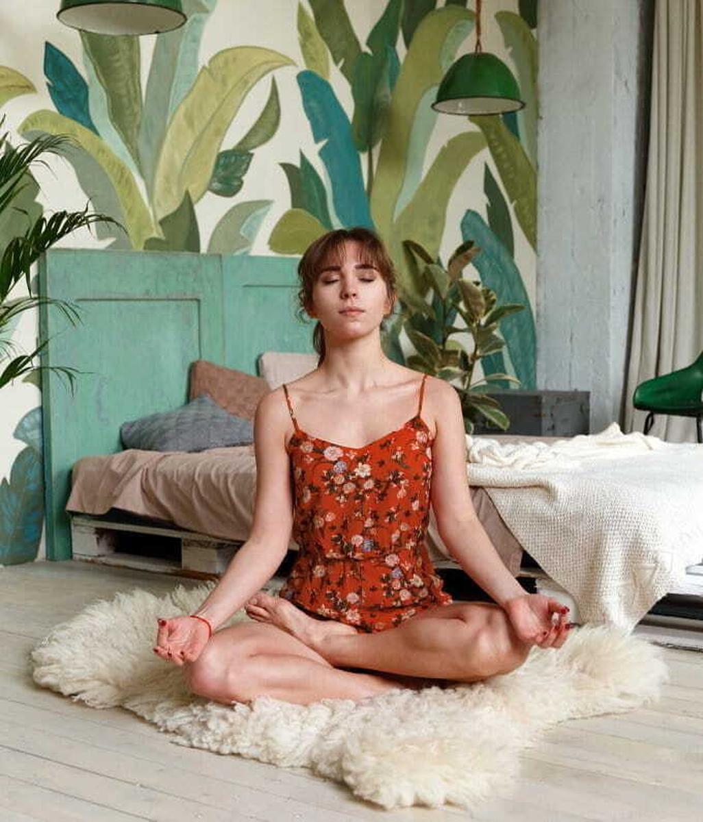 Junge Frau in Kleid in Meditationspose auf dem Boden vor einem Bett aus Paletten und einer mit Pflanzen bemalten Wand