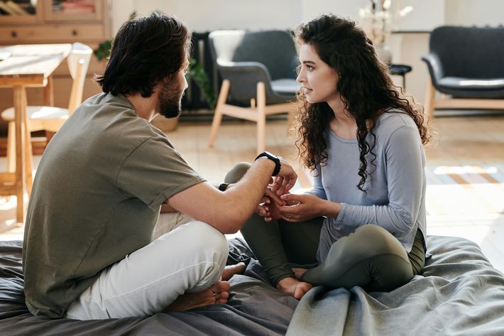 Ein Mann und eine Frau sitzen sich barfuß auf einem Bett gegenüber und führen ein intensives Gespräch. Beide halten sich an den Händen und schauen sich aufmerksam an. Die Umgebung wirkt gemütlich und hell, mit Holzmöbeln und weichen Farben im Hintergrund.