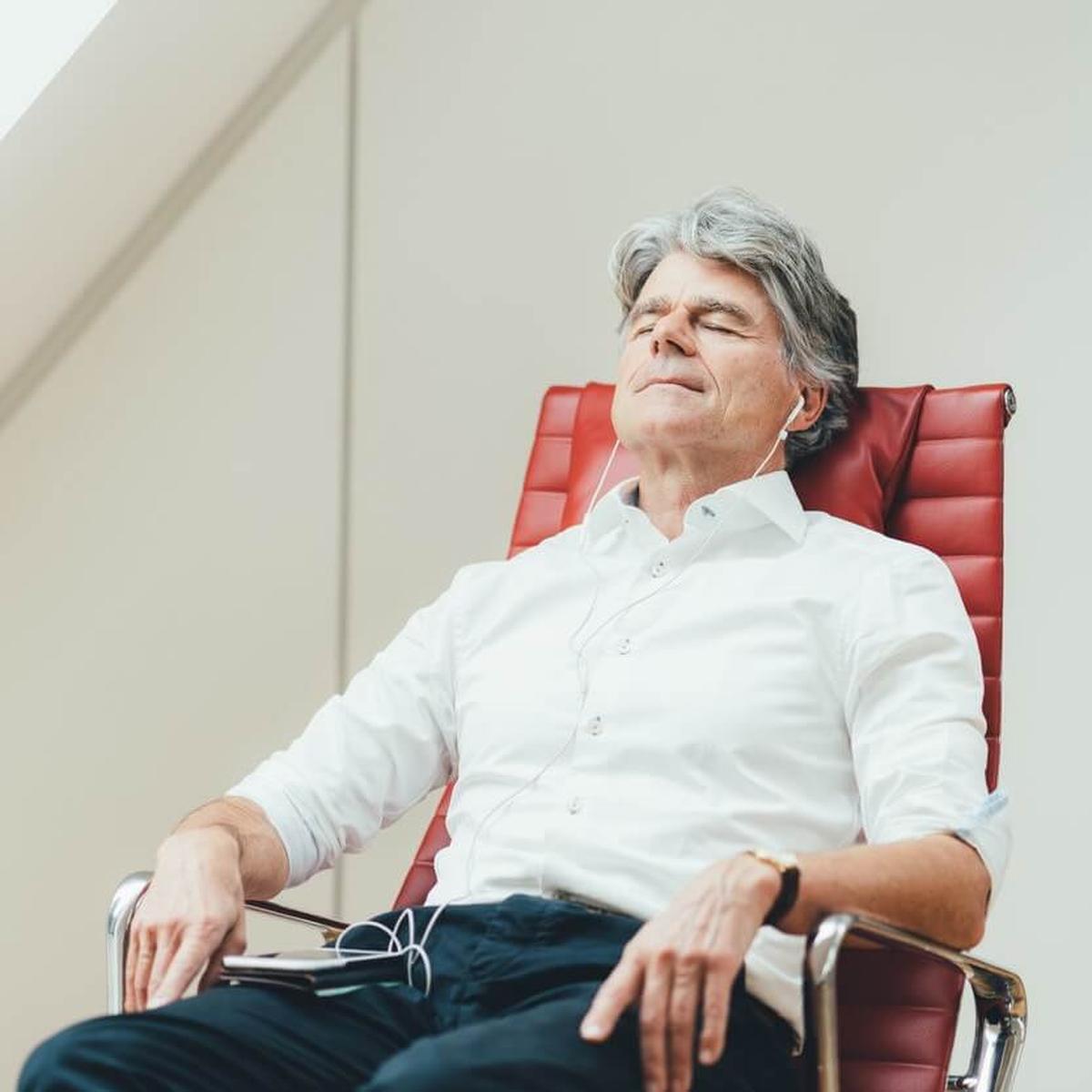 Älterer Mann in business casual Outfit sitzt in rotem Lehnstuhl mit geschlossenen Augen und meditiert mit 7Mind. Quadratisches Bild.
