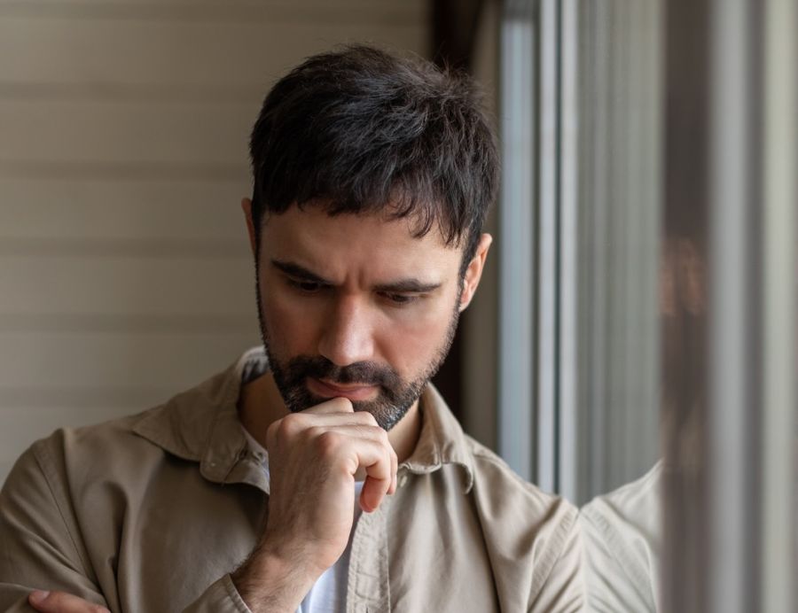 Ein Mann mittleren Alters mit dunklem Haar und Bart steht nachdenklich am Fenster. Er trägt ein beiges Hemd und stützt sein Kinn auf die Hand, während er mit gesenktem Blick und konzentrierter Miene zu grübeln scheint. Das weiche Tageslicht fällt durch die Fenster und beleuchtet sein Gesicht, wodurch eine ruhige, kontemplative Atmosphäre entsteht.