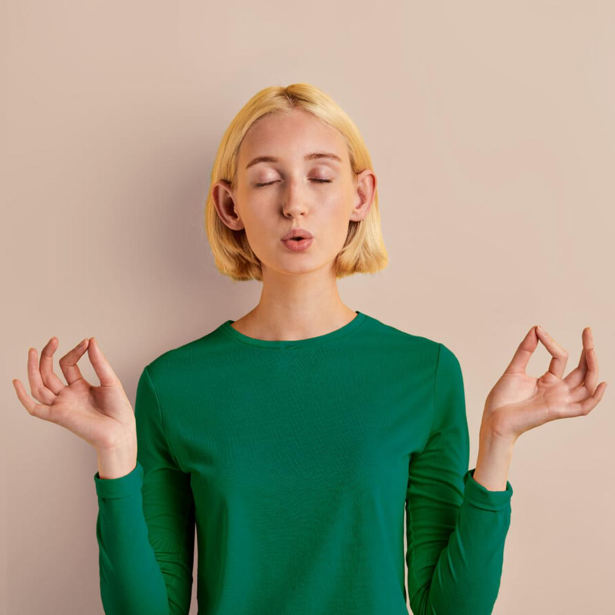 Junge Frau mit kurzen blonden Haaren und grünem T-Shirt hat die Augen geschlossen, den Mund geöffnet und hält an beiden Händen Daumen und Zeigefinger aneinander. Quadratisches Format.