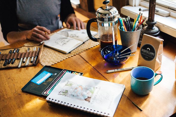 Auf einem Holztisch liegen zwei Skizzenblöcke, Stifte, eine Frenchpress und eine Kaffeetasse und vor der einen Skizze sitzt eine Person mit Bleistift in der Hand.