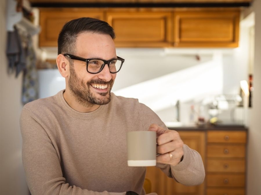 A man with glasses in a beige sweater is sitting in a bright kitchen, holding a mug with one hand. The background shows wooden cabinets and sunlight coming in through a window.