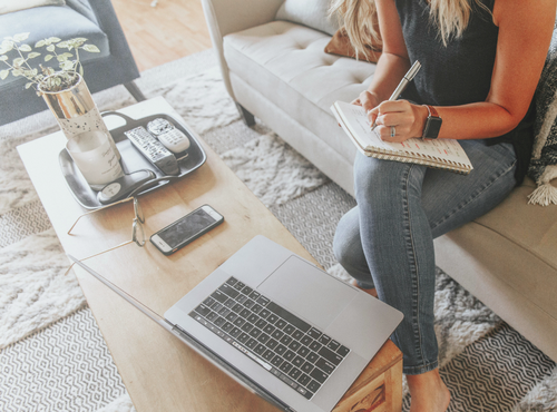 Eine Person sitzt auf einem Sofa und schreibt in ein Notizbuch. Vor ihr auf dem Couchtisch stehen ein aufgeklappter Laptop, ein Smartphone und eine kleine Pflanze.