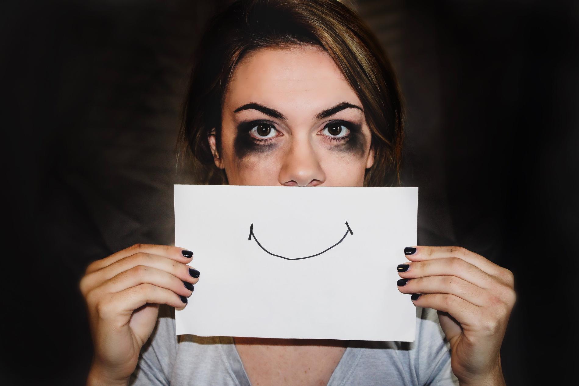 Eine Frau mit verschmiertem Augen Make-Up, braunen Augen, braunen Haaren und schwarz lackierten Fingernägeln schaut frontal in die Kamera und hält ein Papier mit einem aufgezeichneten, lächelnden Mund vor ihren Mund.