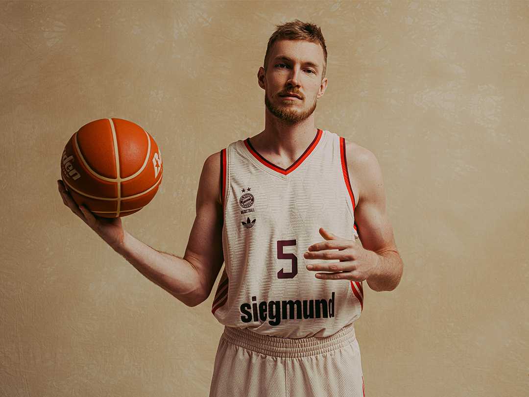 Basketball player in a white jersey with red trim holding a basketball against a beige background.