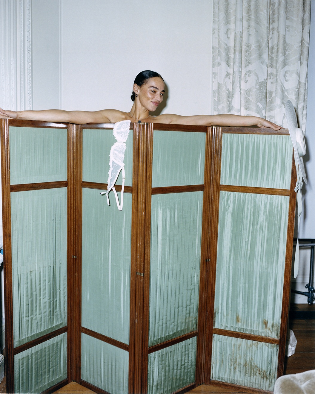 Woman smiling behind a folding screen with lingerie hanging, ornate curtains in the background.