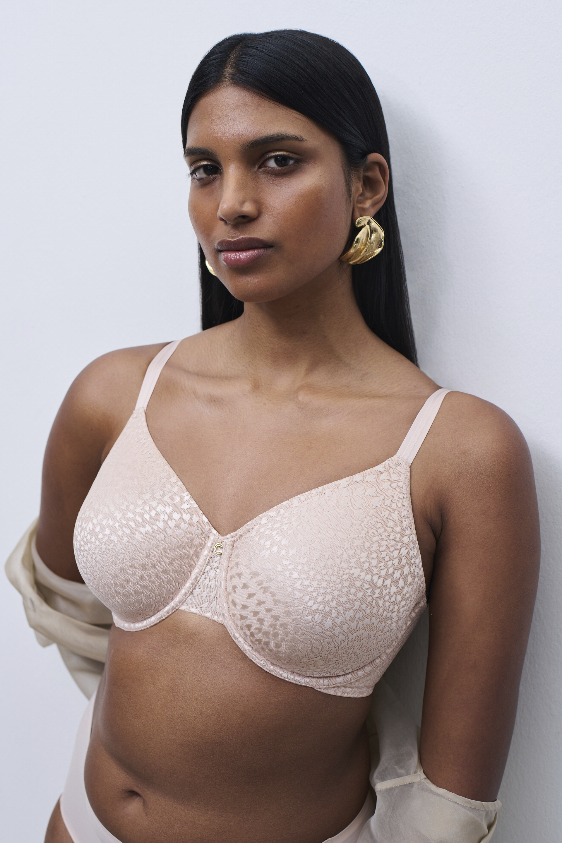 A woman with long dark hair wears a beige patterned bra and gold earrings, standing against a white background.