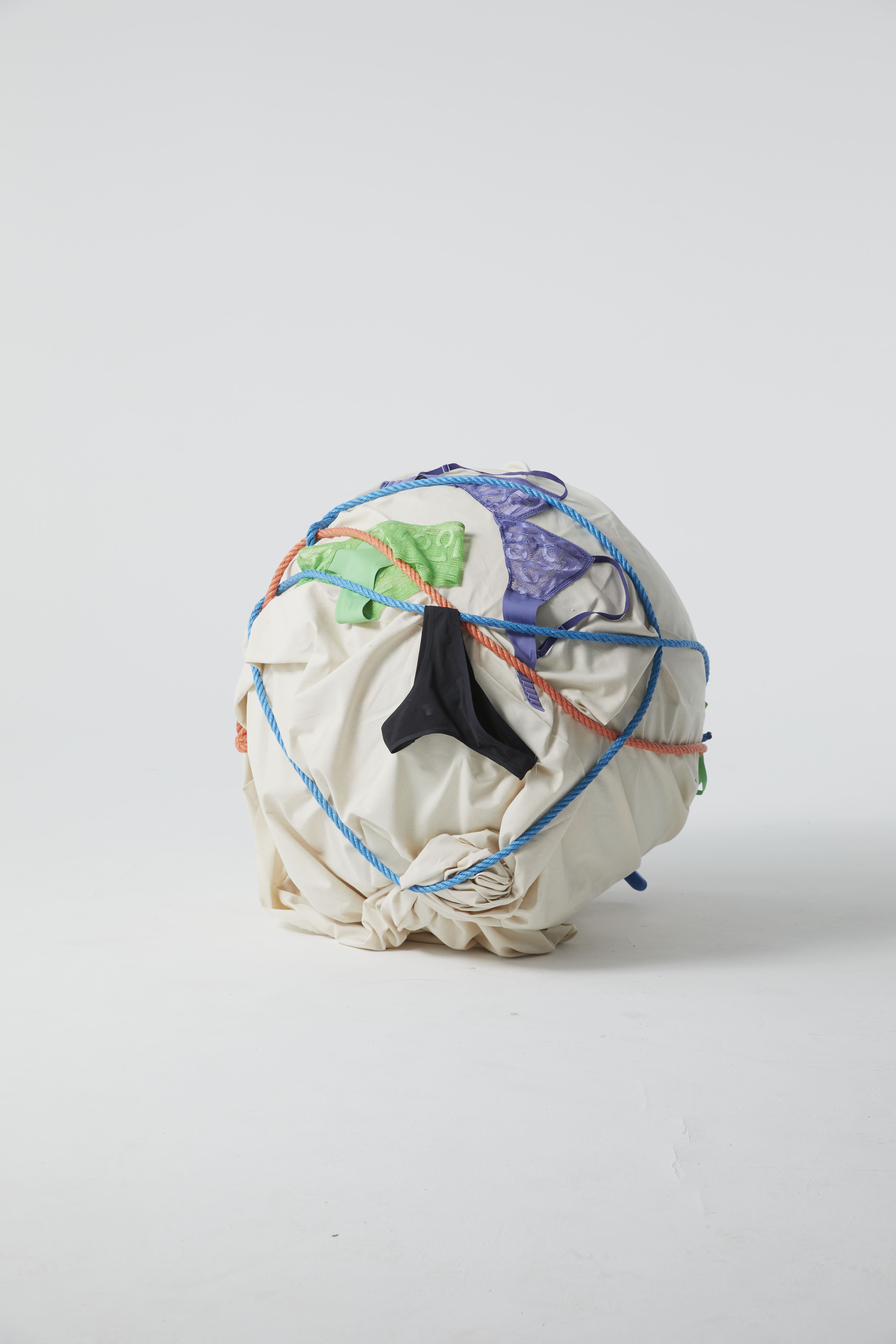 A large, round object wrapped in beige fabric and colorful ropes, with a black plastic piece visible among the bindings.