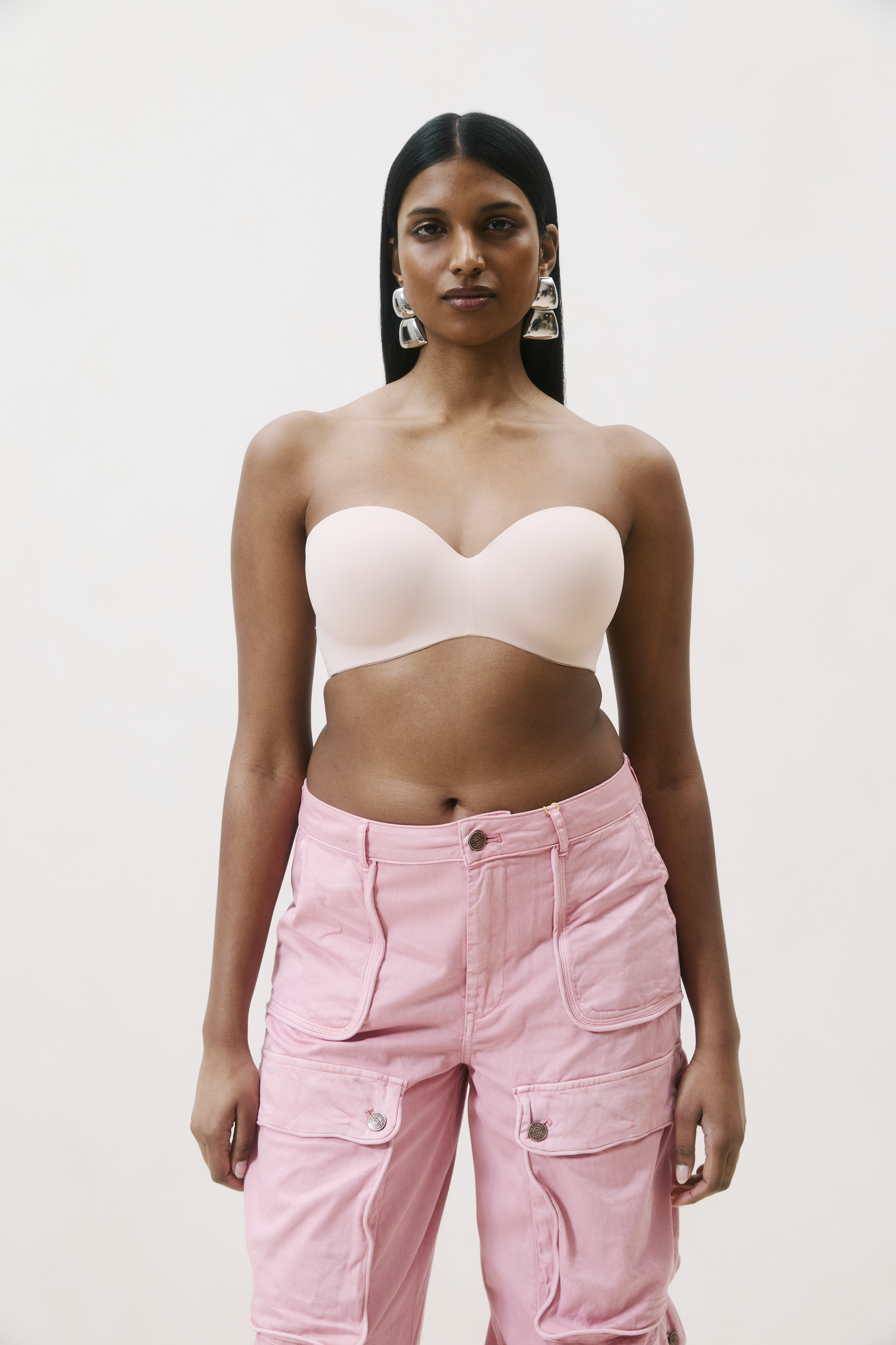 A person stands against a plain background wearing a nude strapless top and pink cargo pants, with long straight hair and large hoop earrings.
