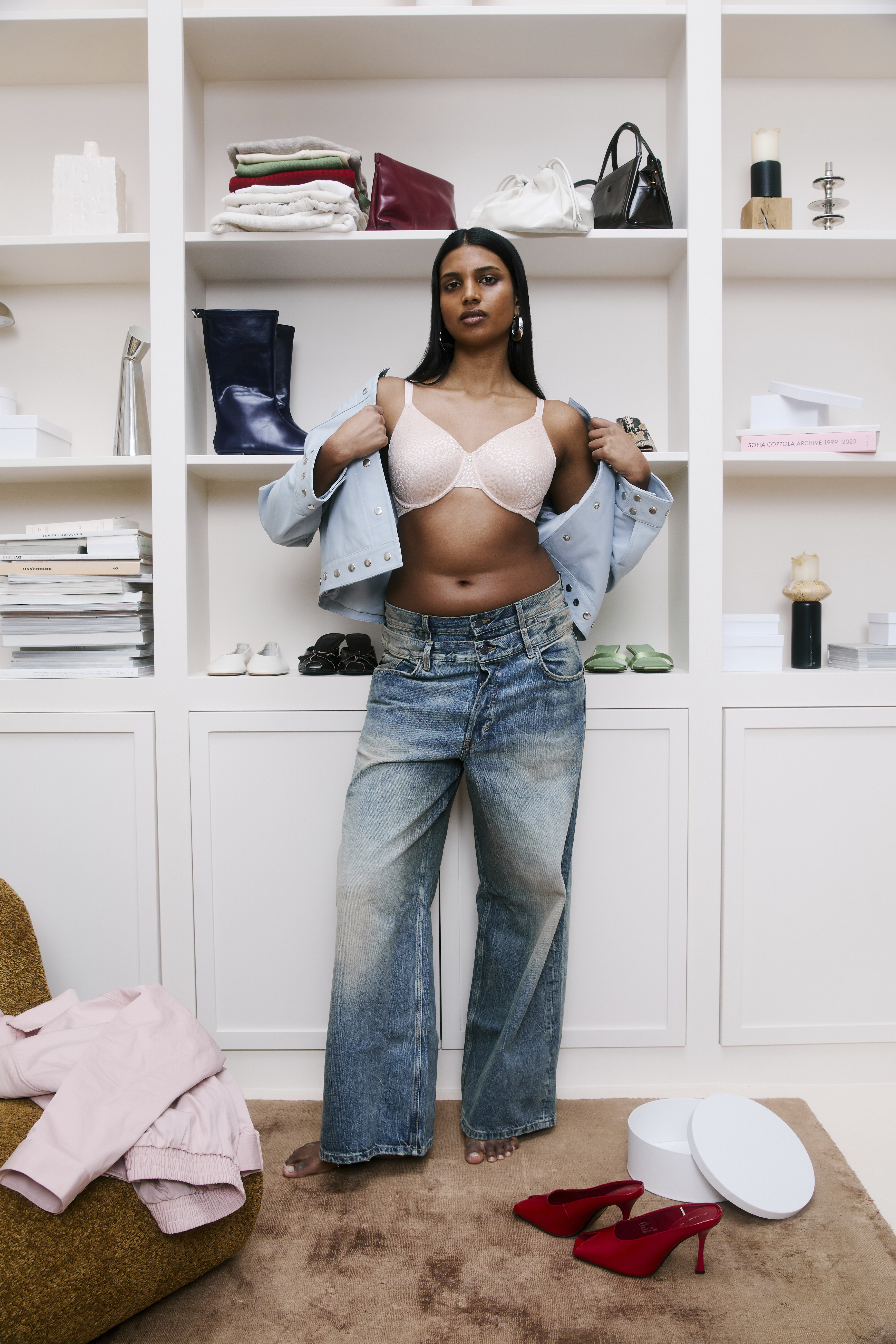 Woman in jeans and a bra tries on a jacket in a chic wardrobe with shelves of bags and books, with red heels and clothes nearby.