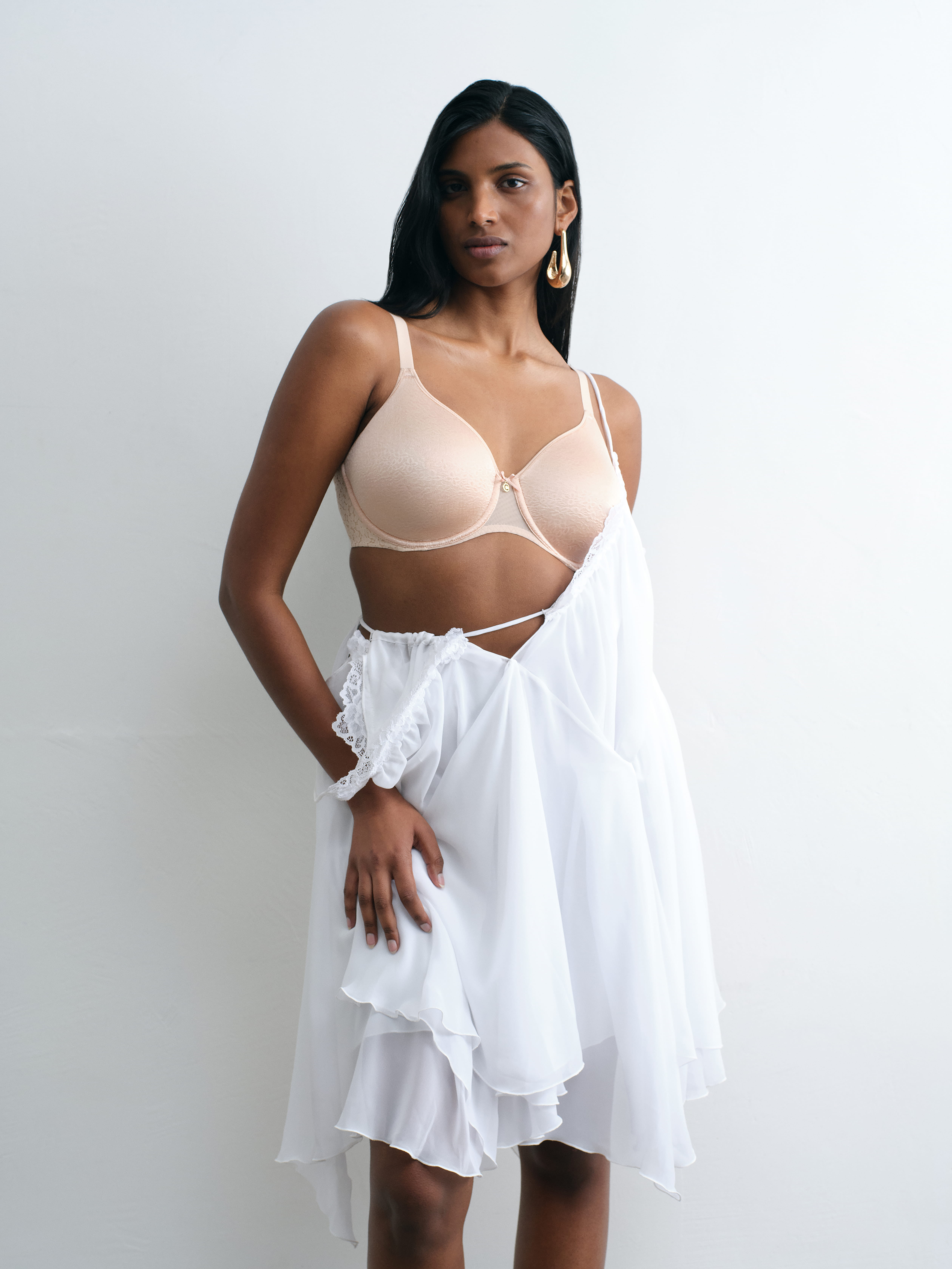 A person wearing a beige bra and a flowing white skirt, standing against a plain white background.
